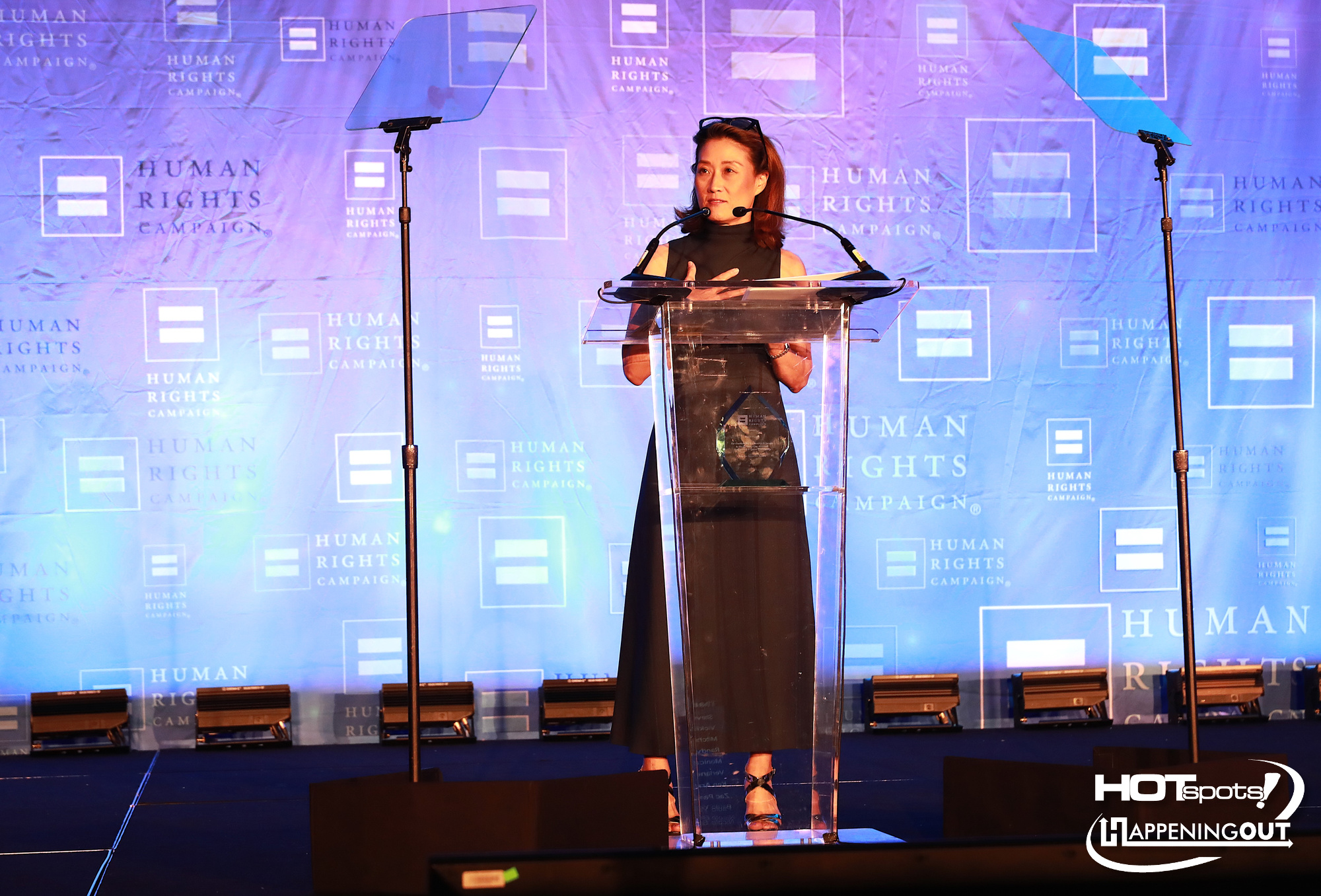 Woman speaker at a clear acrylic podium giving a talk at a Human Rights Campaign event, blue branded backdrop behind her.