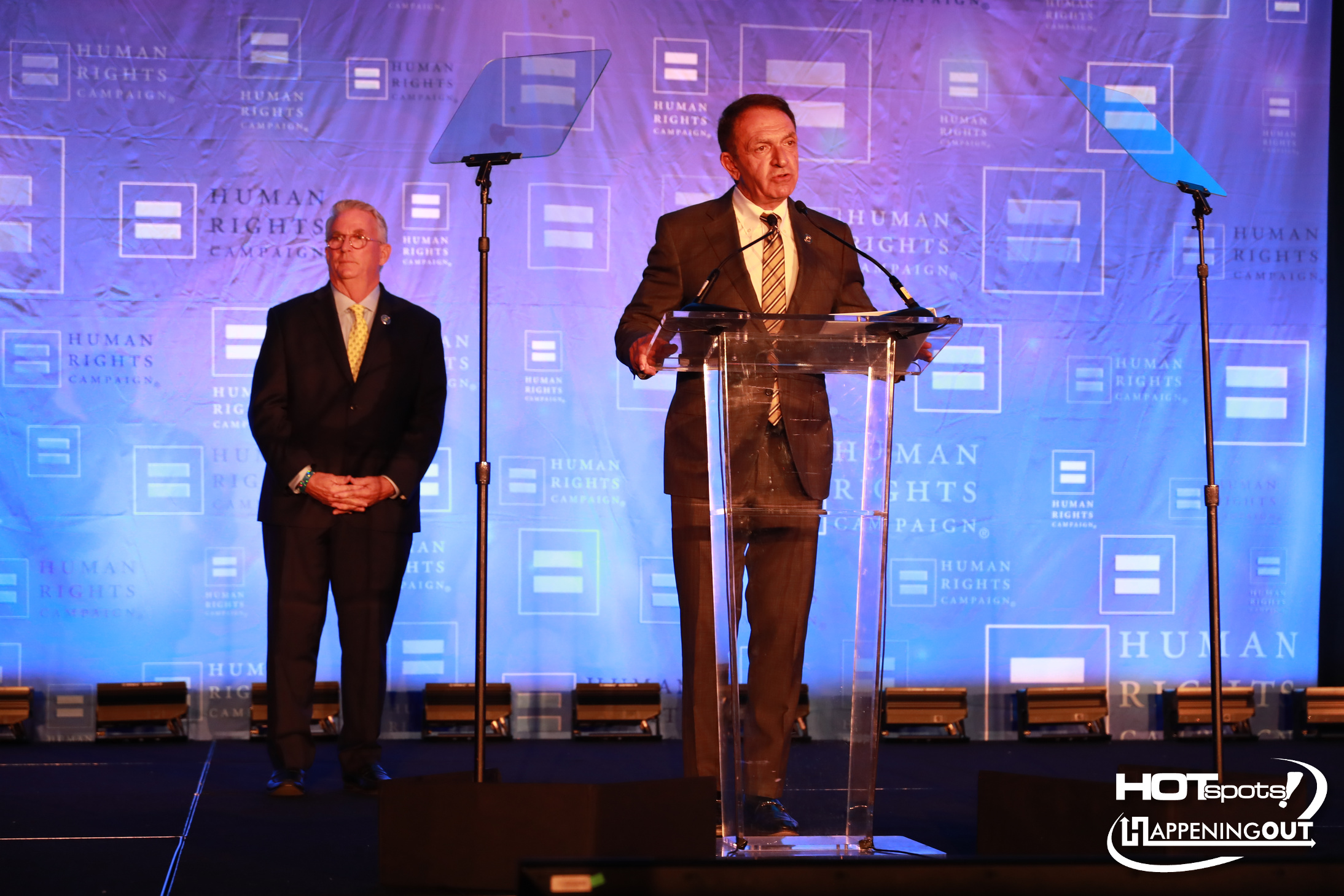 Man in a suit speaks at a clear podium on a stage with a blue Human Rights Campaign backdrop and a second man standing nearby.