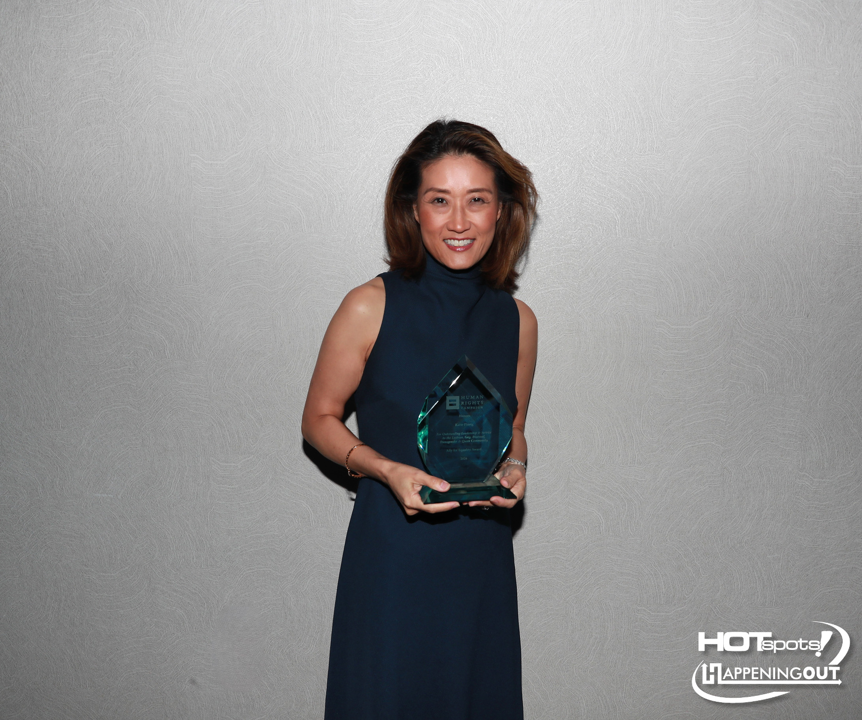 Smiling woman in a navy sleeveless dress holding a crystal award against a textured gray wall; event branding visible in the corner.