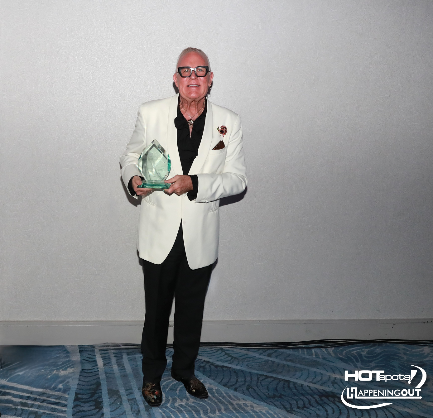 Older man in a white blazer holding a glass award, standing in a hotel/event space.