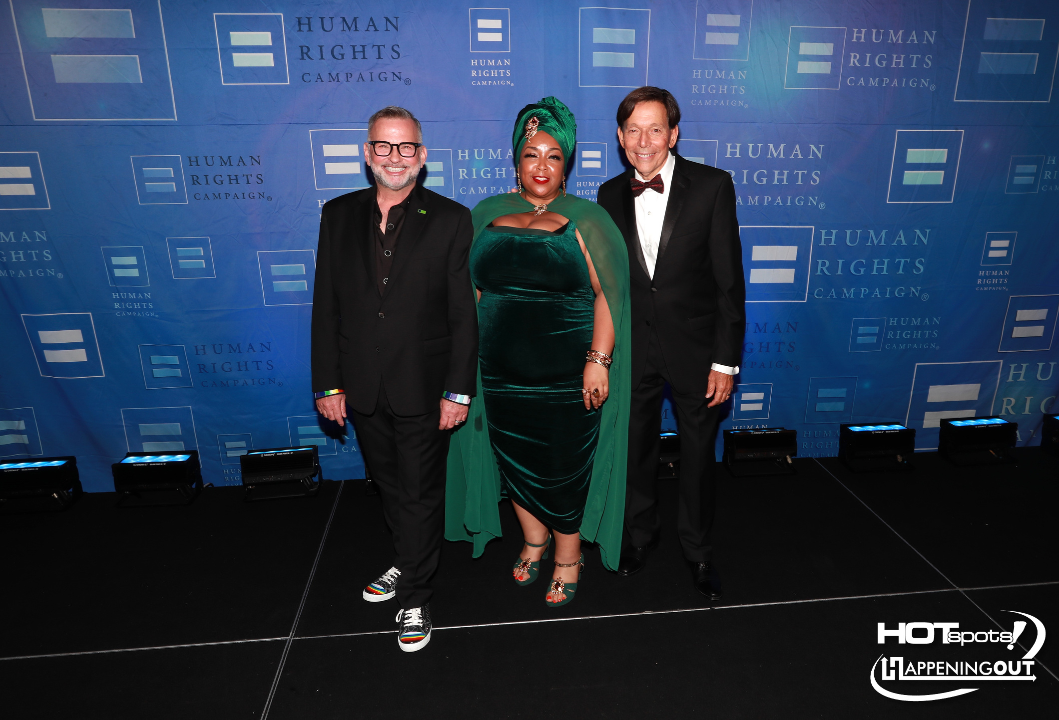 Three people pose on a black carpet in front of a blue Human Rights Campaign backdrop; two men in black suits flank a woman in an emerald green velvet gown with a cape and headwrap, smiling at the camera.