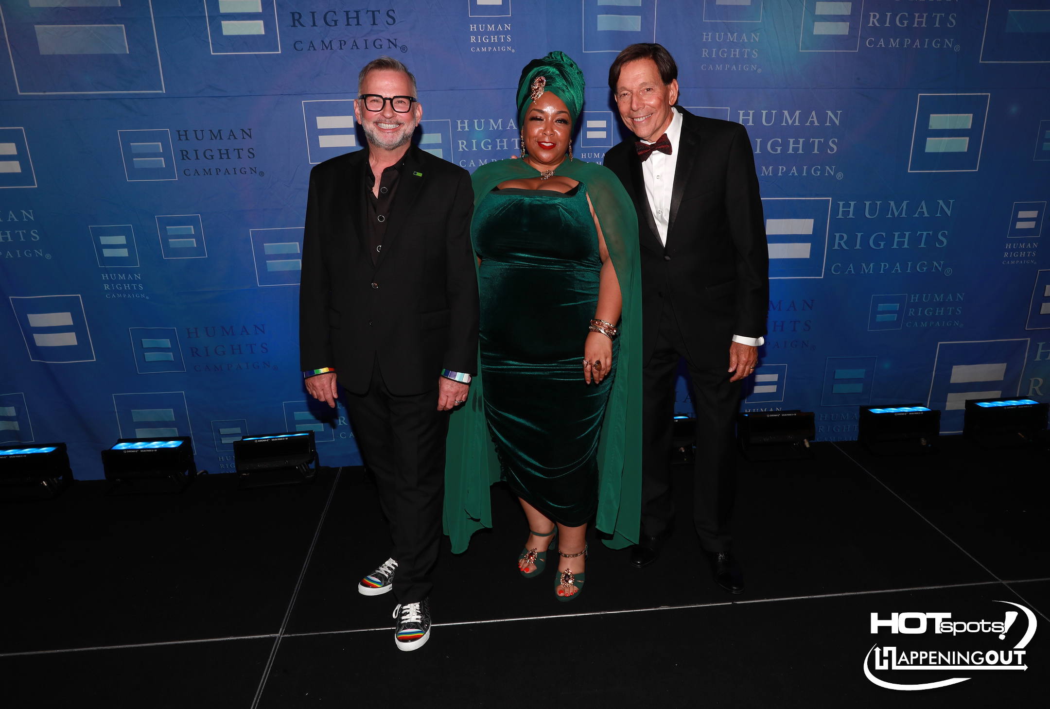 Three people pose together on a blue backdrop at a Human Rights Campaign event: a woman in an emerald velvet gown with a cape and headwrap flanked by two men in black suits, all smiling.