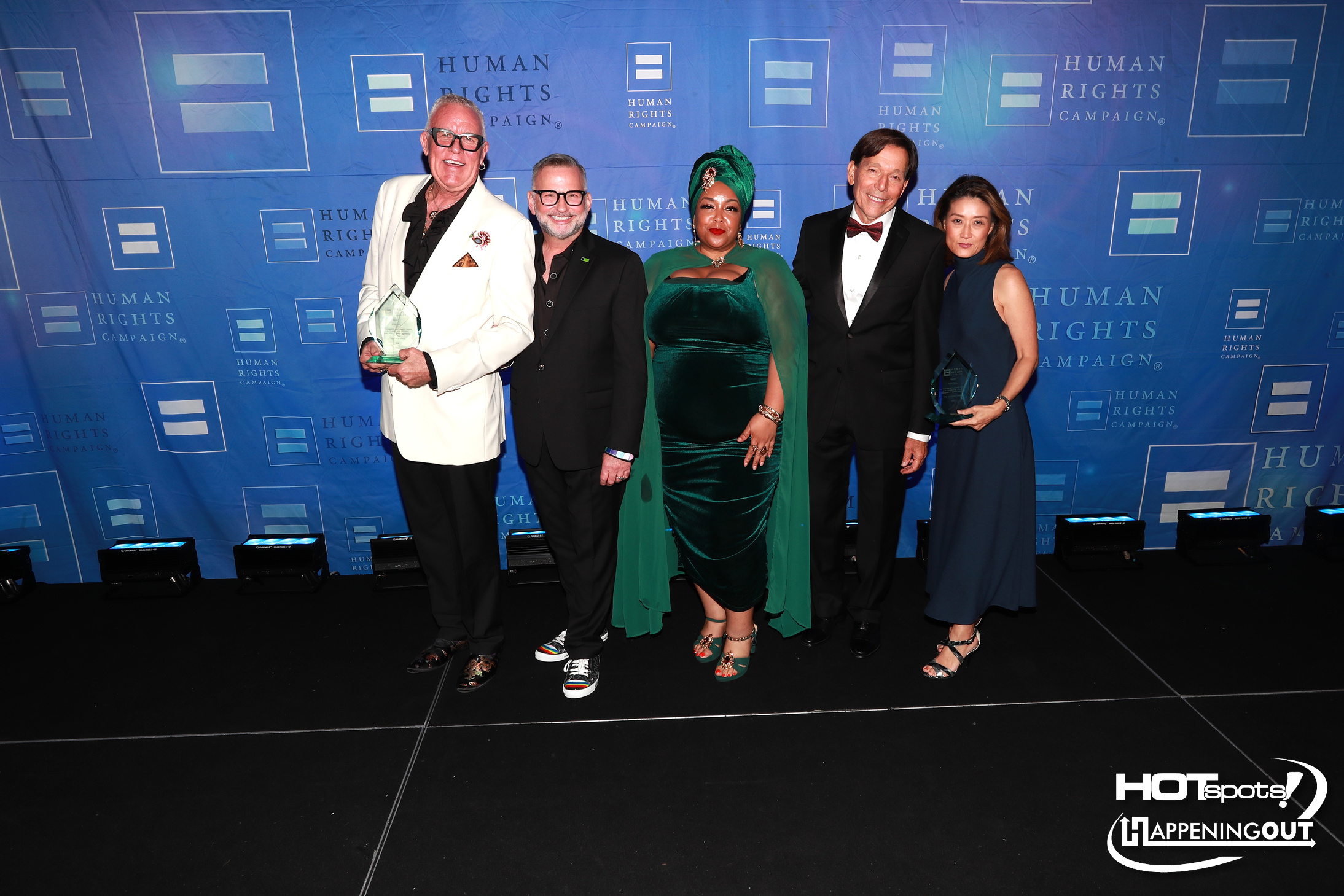 Five people posing on a blue step-and-repeat with Human Rights Campaign logos, each holding or standing near awards at a formal event.