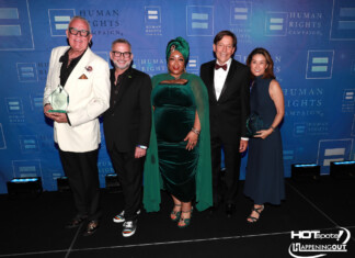 Five adults in formal attire posing together on a stage with a blue Human Rights Campaign backdrop; two men hold clear glass trophies.