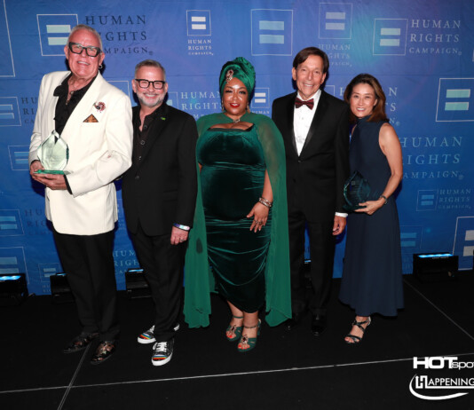 Five adults in formal attire posing together on a stage with a blue Human Rights Campaign backdrop; two men hold clear glass trophies.