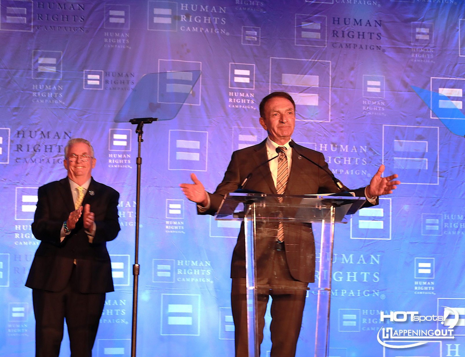 Man at a podium delivering a speech with arms extended, while a second man applauds beside him on a blue backdrop filled with Human Rights Campaign logos.