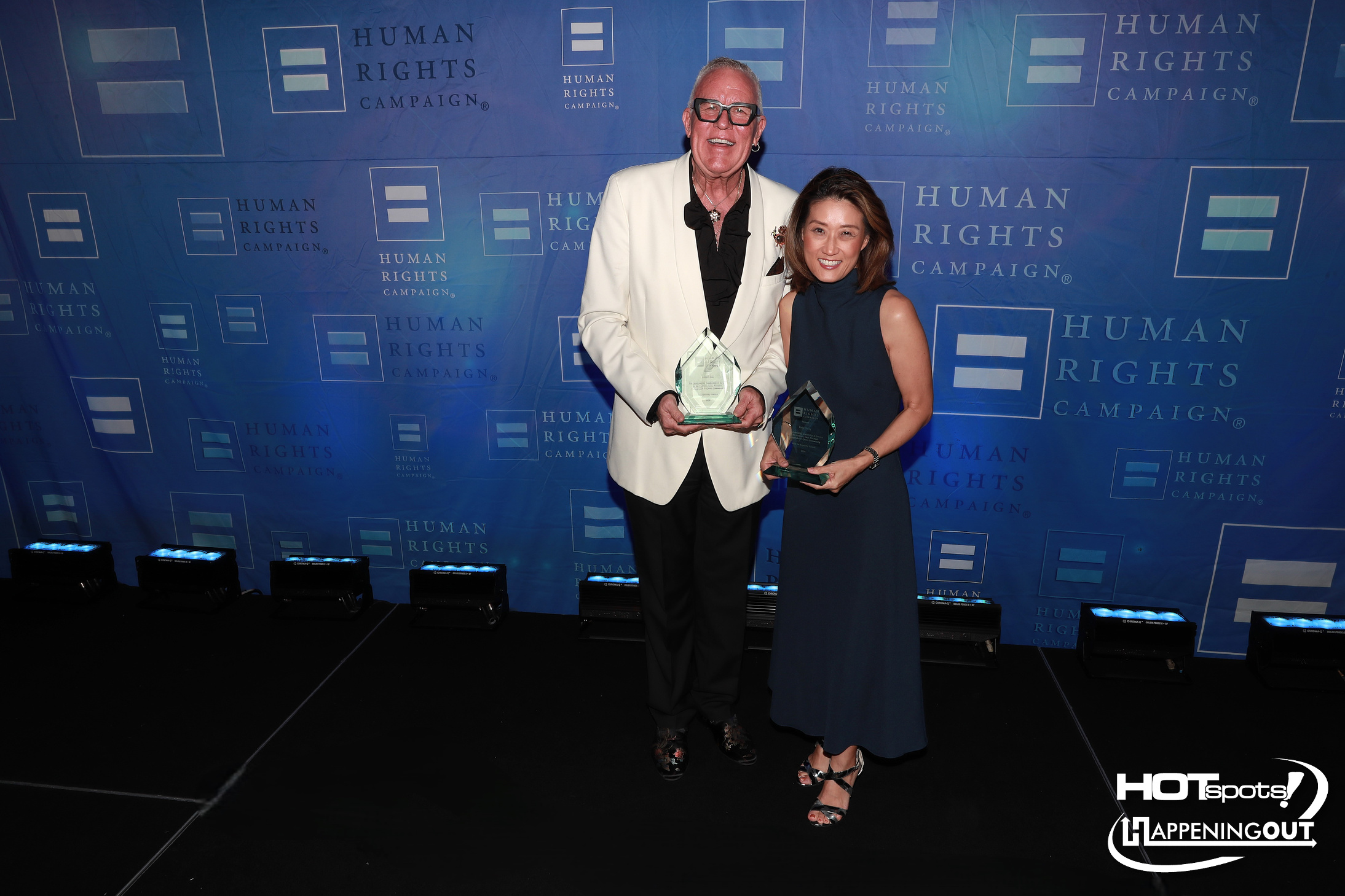 Two award recipients pose with glass trophies at a Human Rights Campaign event; man in a white blazer, woman in a navy sleeveless dress, both smiling.