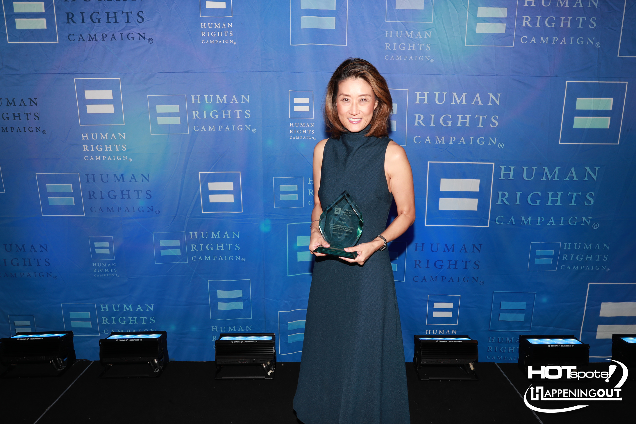 Woman in a sleeveless teal gown on a stage, holding a glass award, with a blue backdrop featuring Human Rights Campaign logos.
