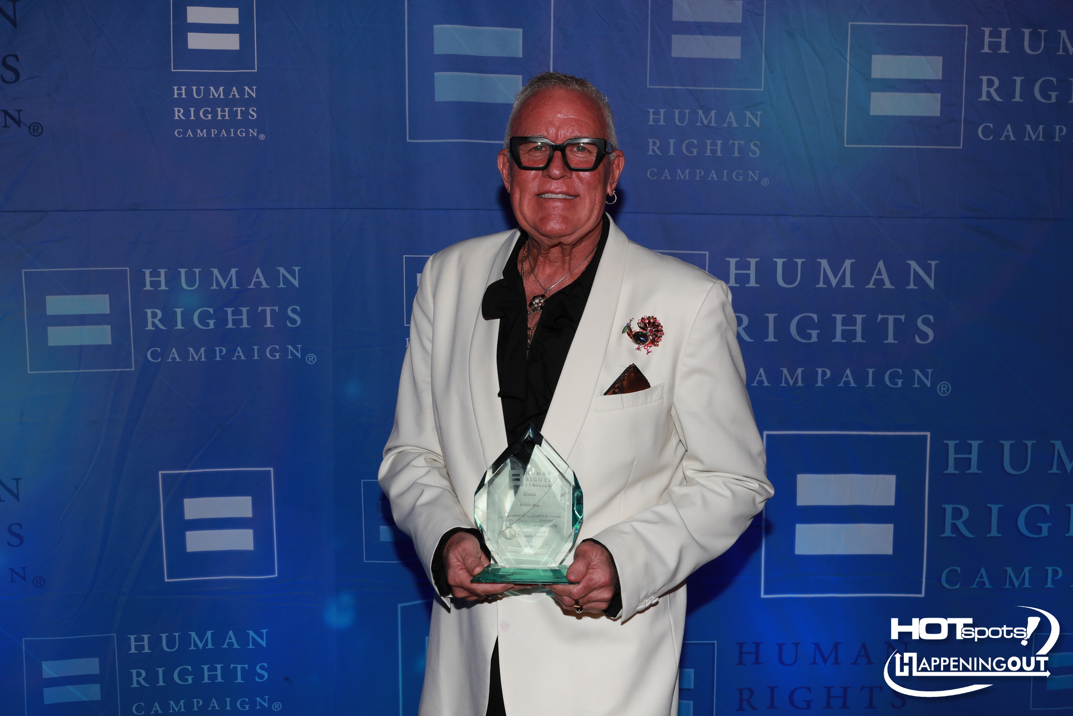 Man in a white blazer and black shirt holding a glass trophy at a Human Rights Campaign event, smiling in front of a blue backdrop.