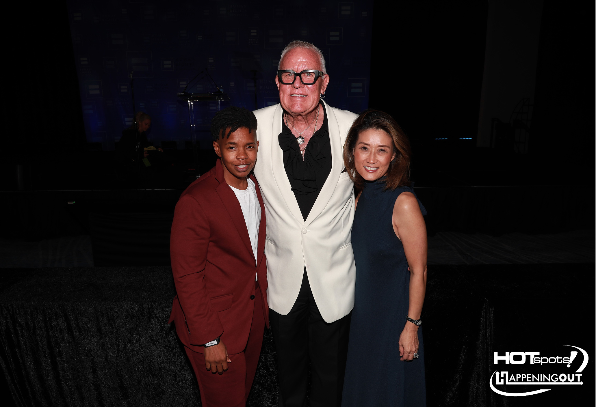 Three people posing together at a formal event: a tall man in a white blazer flanked by a young man in a red suit and a woman in a navy dress, with event backdrop behind them.
