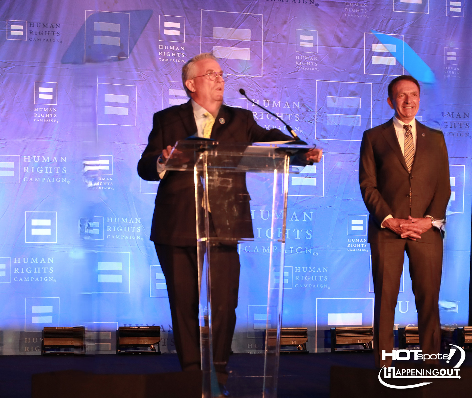 Speaker at a podium on a blue backdrop with Human Rights Campaign logos; another man in a suit stands nearby on stage.