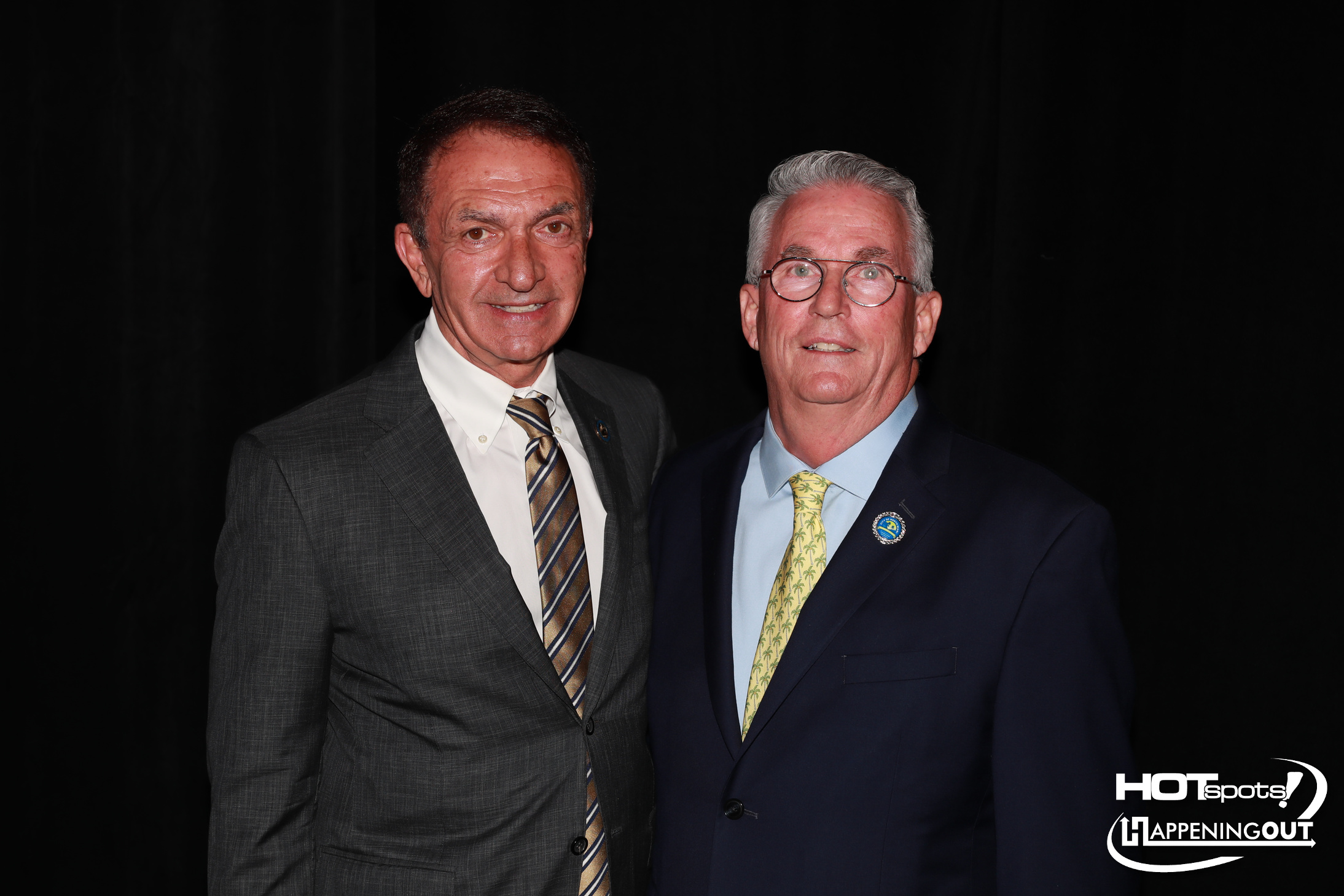 Two men in suits posing for a photo at an event, with a black backdrop and a logo in the corner reading HOTspots Happening Out.