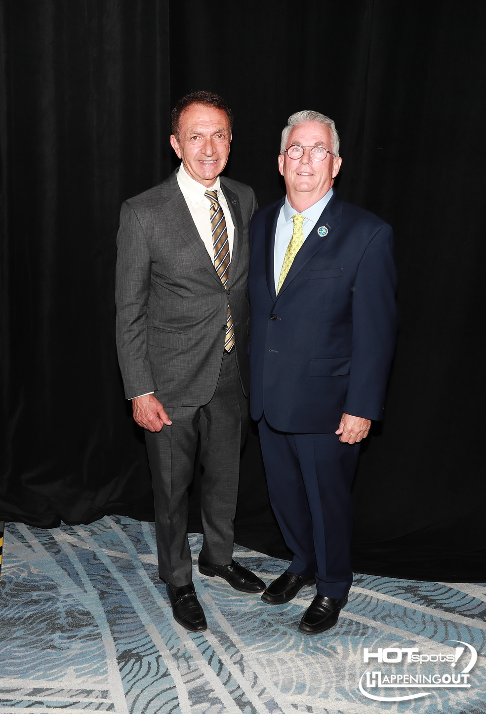 Two men in business suits posing together for a photo at a formal event, standing in front of a black backdrop.