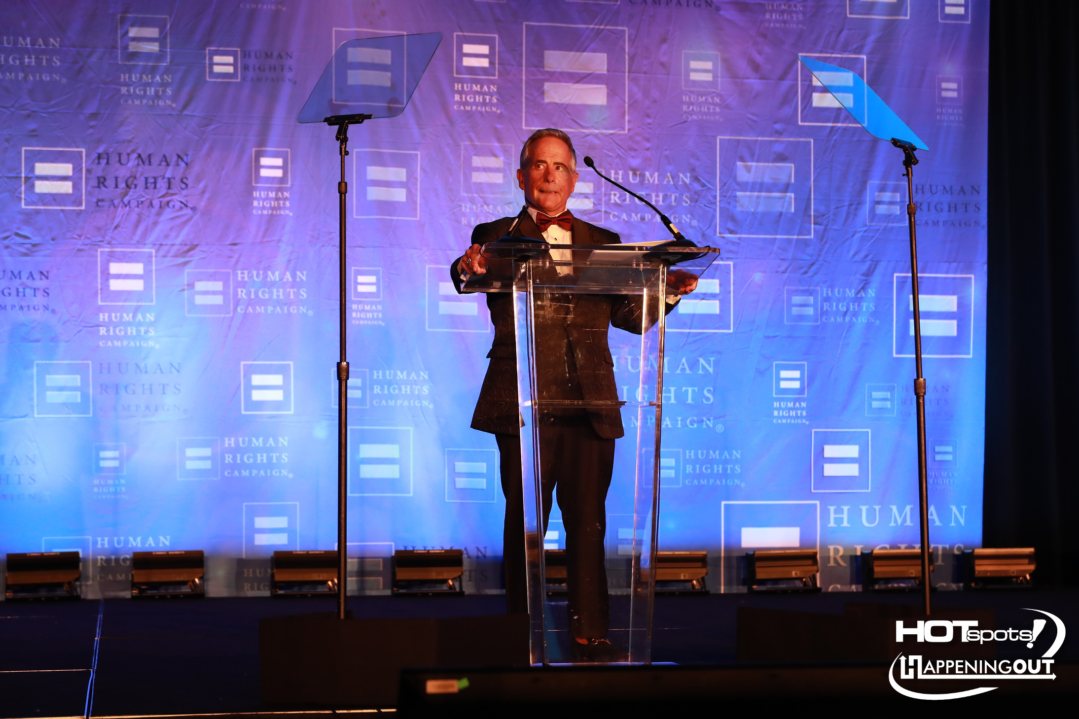 Man in a tuxedo speaks at a clear podium on a stage with a blue backdrop displaying the Human Rights Campaign logo and symbols.