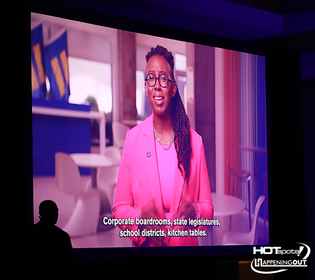 Black woman in a pink blazer speaks on a large screen; captions read: 'Corporate boardrooms, state legislatures, school districts, kitchen tables.' Logo in bottom-right corner.