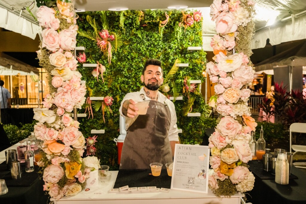 What’s Hot South Florida: April 23rd – May 4th | Hotspots Magazine Hotspots Magazine Bartender in apron extending a drink toward the camera at a floral booth with pink roses and green garden backdrop.