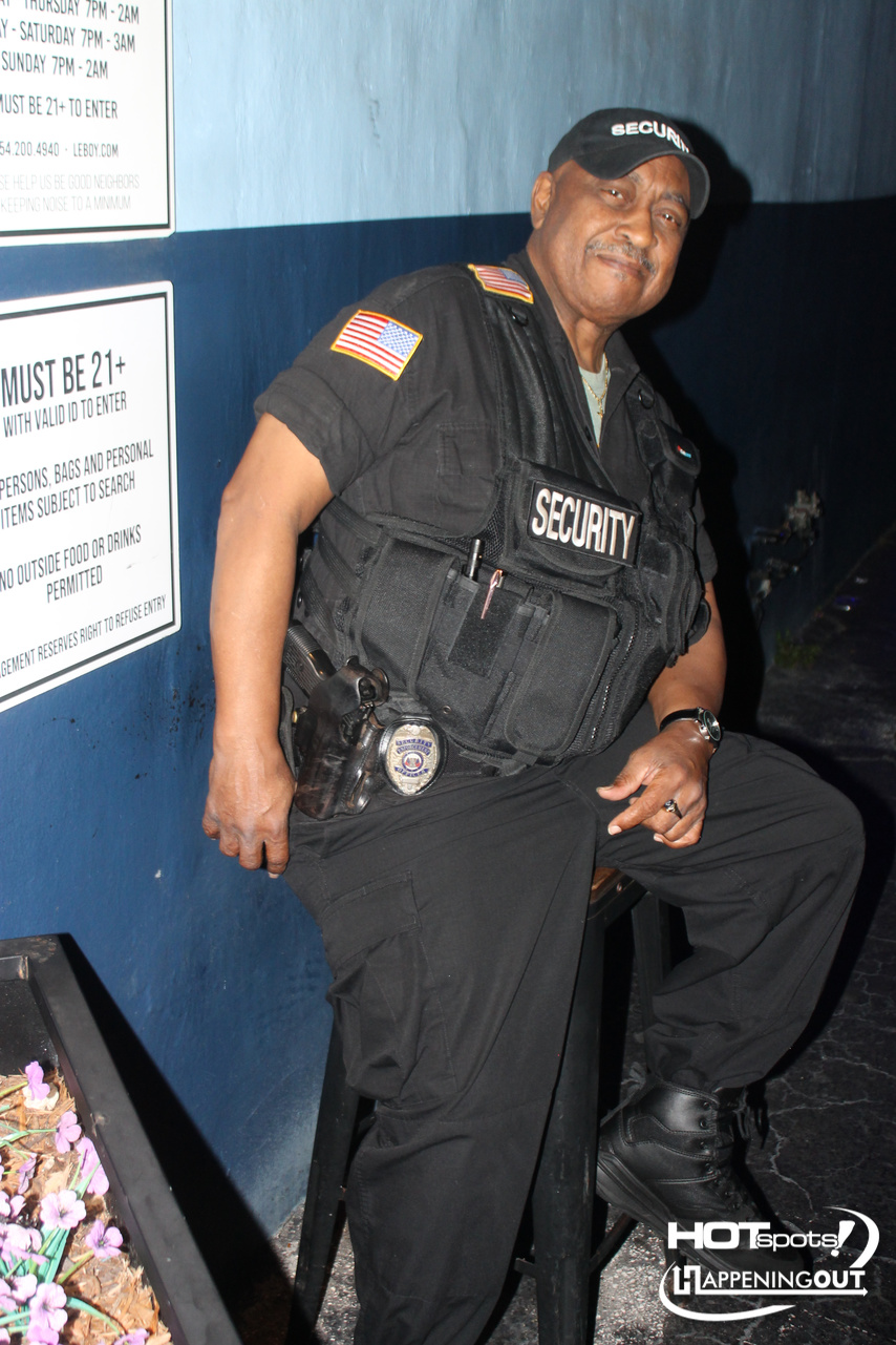 Security guard in a black uniform sits against a blue wall, with a badge, radio, and utility belt visible at his side.