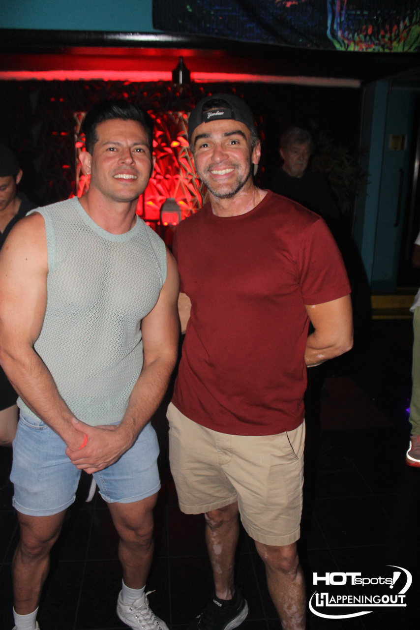 Two men posing and smiling in a nightclub with red lighting; one in a gray sleeveless shirt, the other in a maroon tee and khaki shorts.