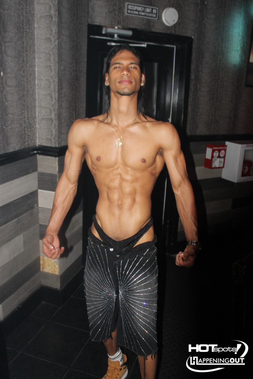 Shirtless man with defined abs posing in a nightclub hallway, wearing glittery pants and a gold necklace.