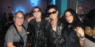 Four friends posing together in a nightclub, wearing dark rock-style outfits and sunglasses, with blue and purple lighting in the background.