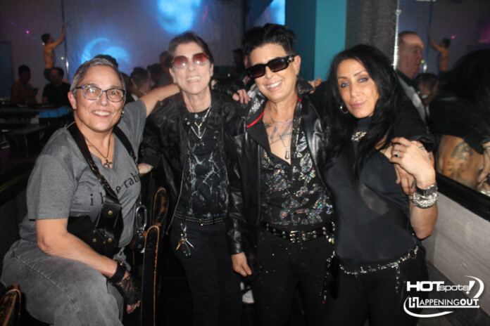 Four friends posing together in a nightclub, wearing dark rock-style outfits and sunglasses, with blue and purple lighting in the background.