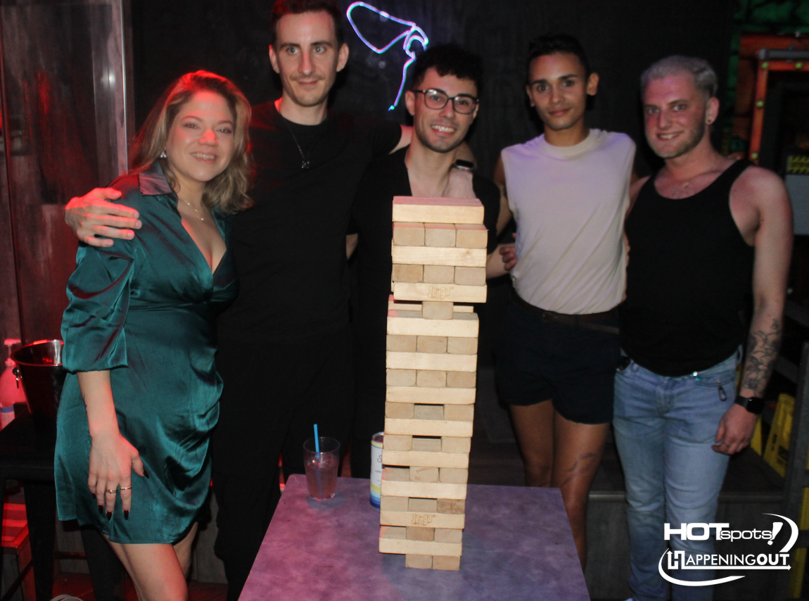 Five friends pose with a tall Jenga tower on a purple table at a party.