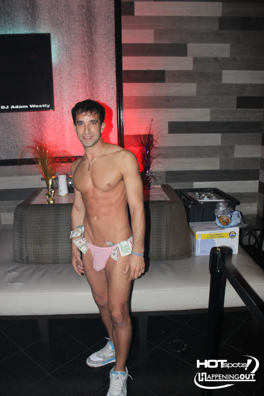 Man posing shirtless in a nightclub wearing pink bikini briefs with dollar bills tucked into the waistband, standing in front of a couch and neon lighting.