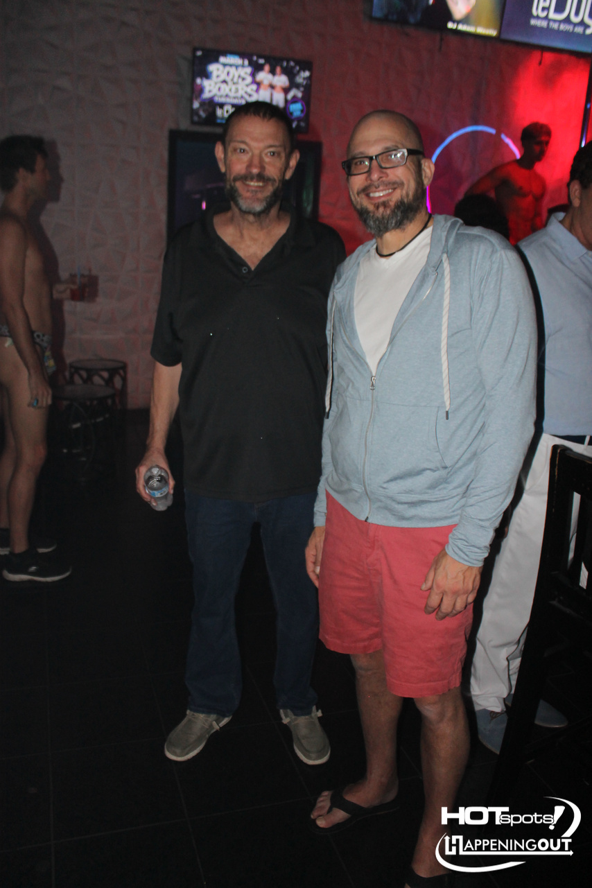 Two smiling men posing for a photo in a nightclub, one in a black polo and dark jeans, the other in a light blue hoodie and red shorts, with red and blue lights in the background.