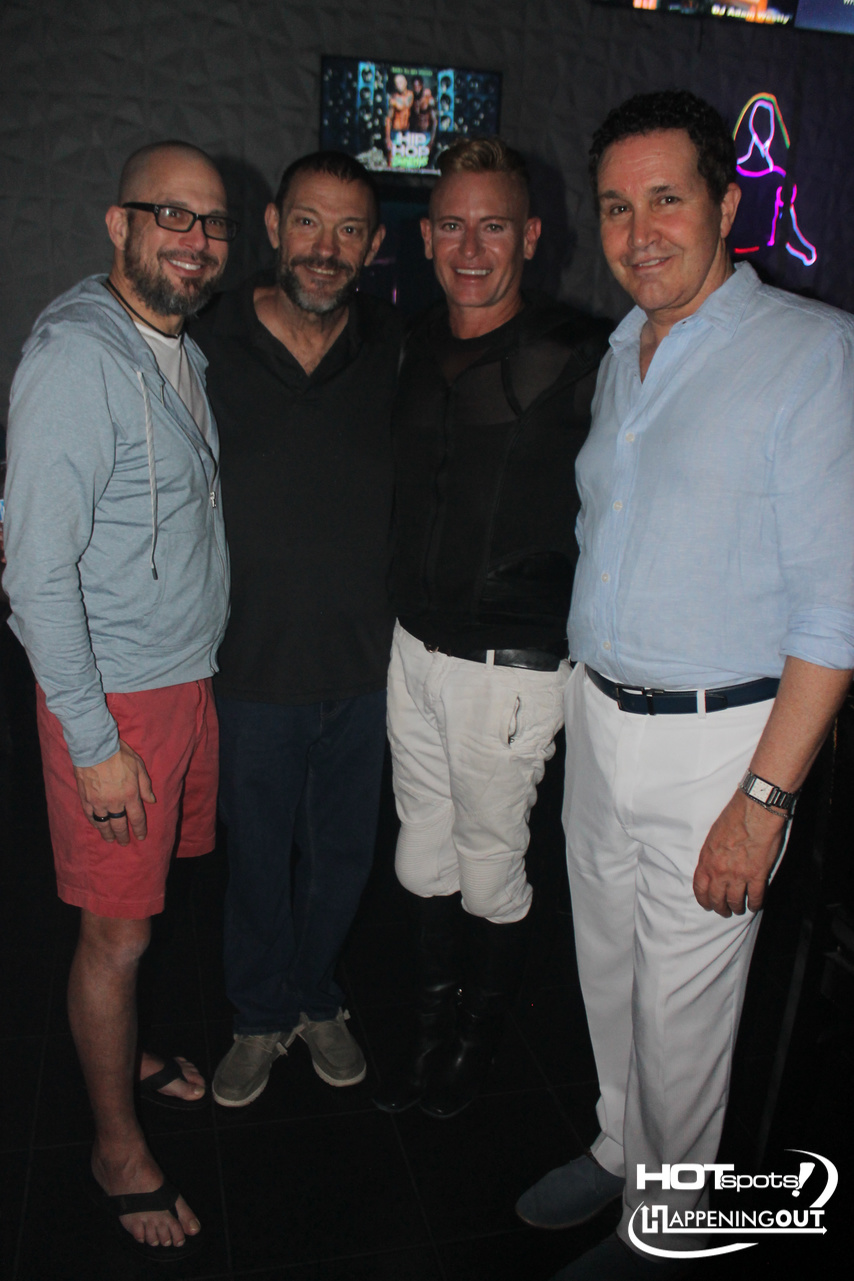 Four men posing together in a nightclub with dim lighting and neon signs in the background.