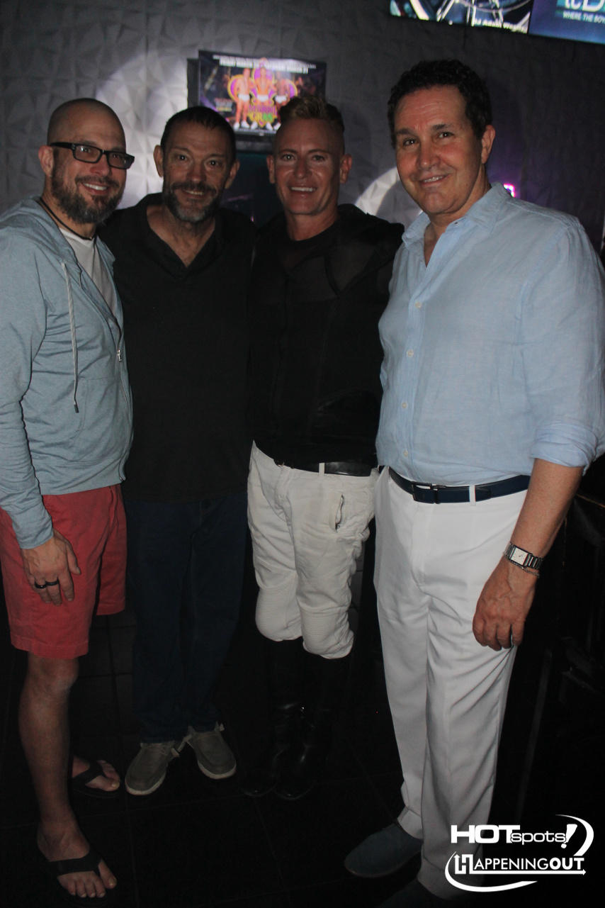 Four men posing together in a dark club, smiling at the camera; one wears a light blue shirt, another in black, with a logo reading 'Hotspots' in the corner.