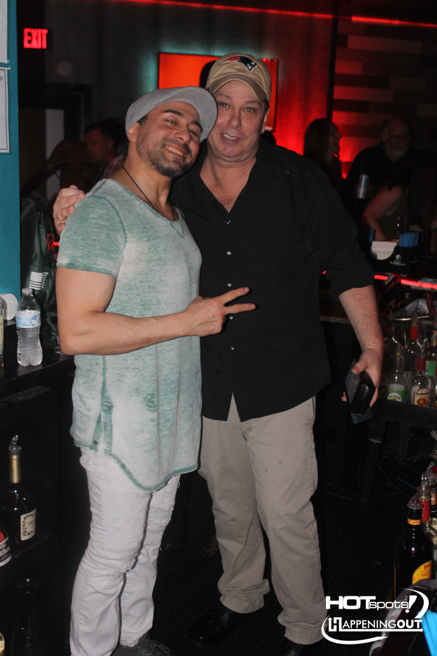 Two men posing for a photo in a bar, smiling; one wearing a gray hat and mint T-shirt, the other in a black shirt and beige pants.