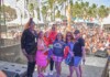 Six diverse adults pose on an outdoor stage at a Pride festival, with a crowded audience, tents, rainbow flags, and palm trees in the background.
