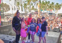 Six diverse adults pose on an outdoor stage at a Pride festival, with a crowded audience, tents, rainbow flags, and palm trees in the background.