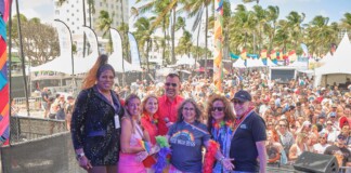 Six diverse adults pose on an outdoor stage at a Pride festival, with a crowded audience, tents, rainbow flags, and palm trees in the background.