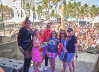 Six diverse adults pose on an outdoor stage at a Pride festival, with a crowded audience, tents, rainbow flags, and palm trees in the background.