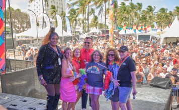 Six diverse adults pose on an outdoor stage at a Pride festival, with a crowded audience, tents, rainbow flags, and palm trees in the background.