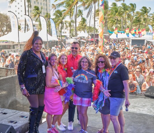 Six diverse adults pose on an outdoor stage at a Pride festival, with a crowded audience, tents, rainbow flags, and palm trees in the background.