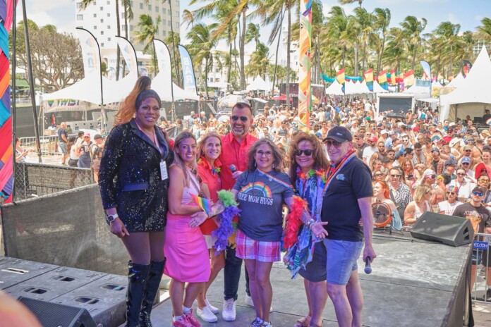 Six diverse adults pose on an outdoor stage at a Pride festival, with a crowded audience, tents, rainbow flags, and palm trees in the background.