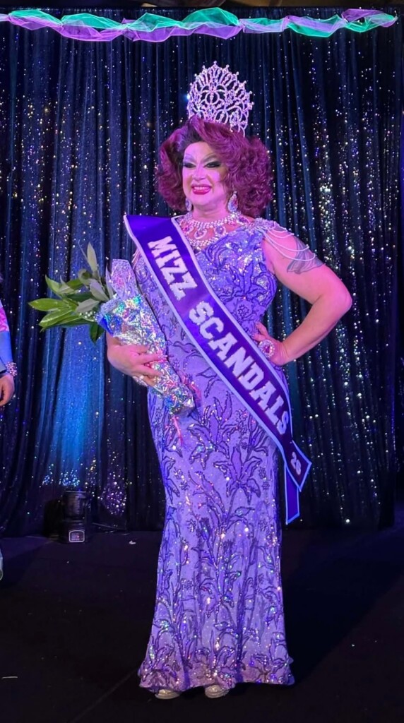 Drag queen winner on stage wearing a purple gown, a jeweled crown, and a Miss Canada sash, holding a bouquet of flowers.