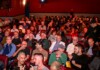 Large audience seated in red theater chairs in a dim auditorium, facing the stage/ screen.