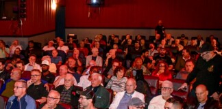 Large audience seated in red theater chairs in a dim auditorium, facing the stage/ screen.