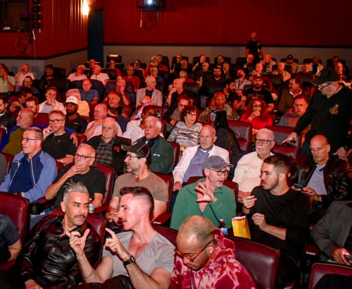Large audience seated in red theater chairs in a dim auditorium, facing the stage/ screen.