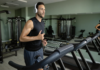 Man in a black sleeveless shirt with white headphones runs on a treadmill in a modern gym with weights and machines in the background.