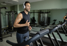 Man in a black sleeveless shirt with white headphones runs on a treadmill in a modern gym with weights and machines in the background.