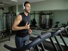 Man in a black sleeveless shirt with white headphones runs on a treadmill in a modern gym with weights and machines in the background.