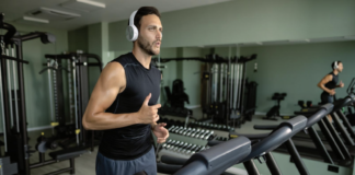 Man in a black sleeveless shirt with white headphones runs on a treadmill in a modern gym with weights and machines in the background.