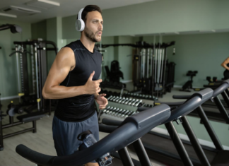 Man in a black sleeveless shirt with white headphones runs on a treadmill in a modern gym with weights and machines in the background.