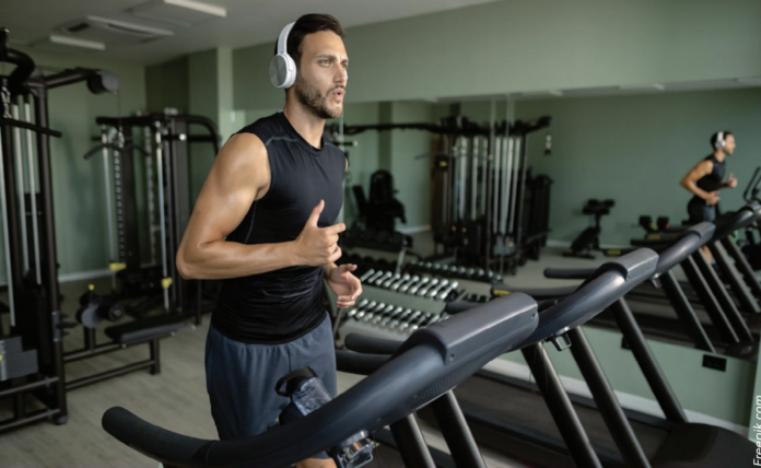 Man in a black sleeveless shirt with white headphones runs on a treadmill in a modern gym with weights and machines in the background.