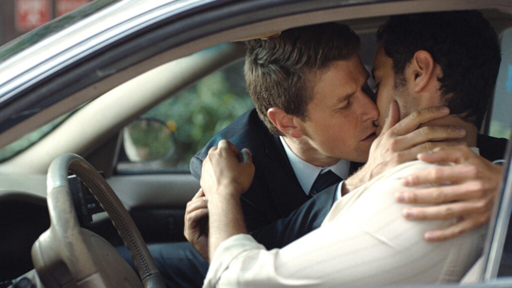 Two men in a car share a kiss, arms wrapped around each other in an intimate hug.