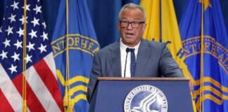 Older man in a suit speaks at a podium with the U.S. flag and department banners behind him.